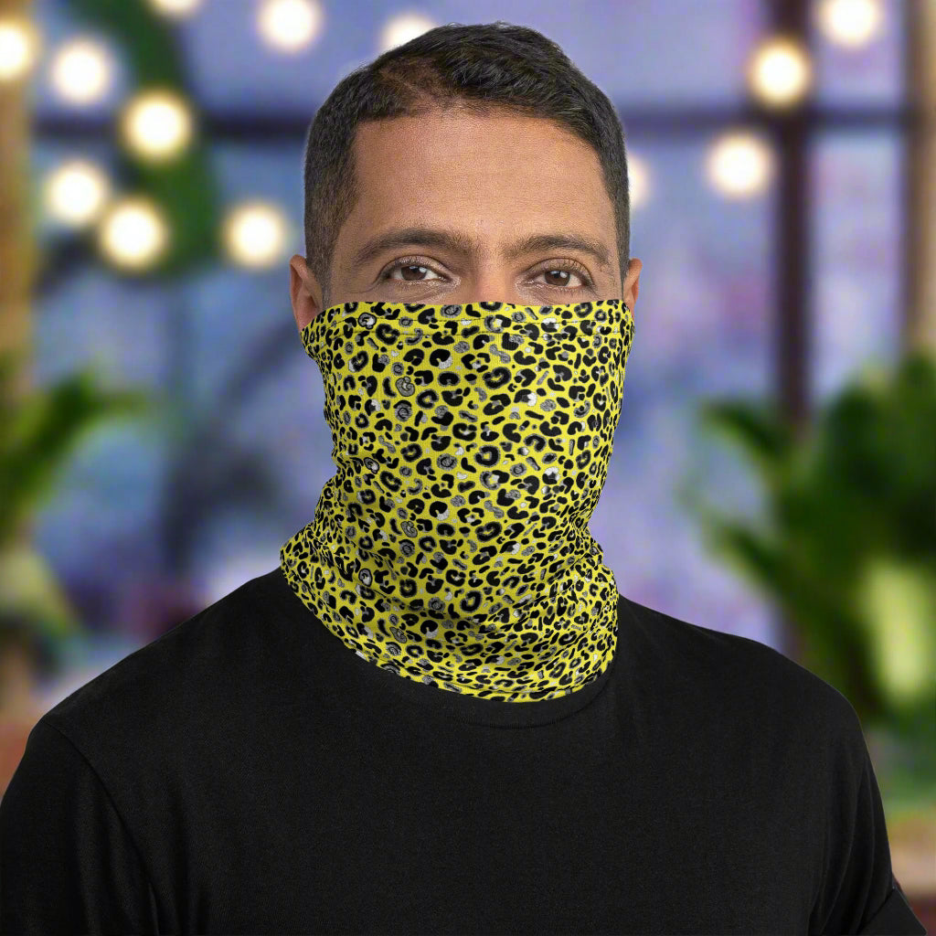 Man with short dark hair facing to the front wearing a black long sleeve top and a vibrant yellow and pink neck gaiter around his neck, covering his nose and mouth. In the background are some fairy lights and plants. This design features an animal print pattern in black, grey and white, printed at a medium scale. The top 3/4 background is bright yellow with the bottom 1/4 a hot pink colour. In this image the pink is not seen.
