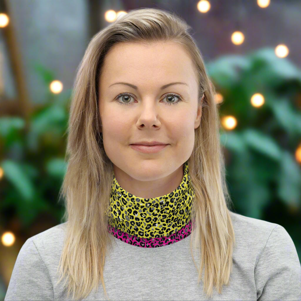 Woman with long blonde hair facing to the front wearing a grey long sleeve top and a vibrant yellow and pink neck gaiter around her neck. In the background are some fairy lights and plants. This design features an animal print pattern in black, grey and white, printed at a medium scale. The top 3/4 background is bright yellow with the bottom 1/4 a hot pink colour