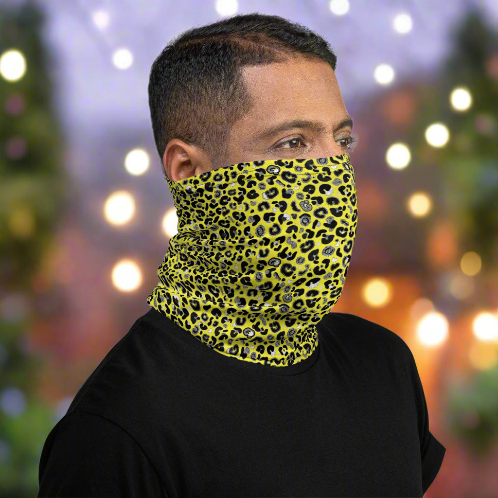 Man with short dark hair facing to the right wearing a black top and a vibrant yellow and pink neck gaiter around his neck, covering his nose and mouth. In the background are some fairy lights and plants. This design features an animal print pattern in black, grey and white, printed at a medium scale. The top 3/4 background is bright yellow with the bottom 1/4 a hot pink colour. In this image the pink is unseen.