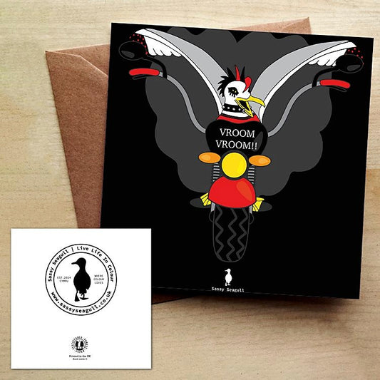 Black Greeting Card with a punk rock seagull with wings spread, riding a motorbike with a cloud of smoke behind it.  The card is on a brown envelope on a wooden table.