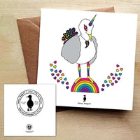 White greeting card with a rainbow unicorn seagull standing on a rainbow with rainbow coloured hearts. Card is on a brown envelope on a brown table.