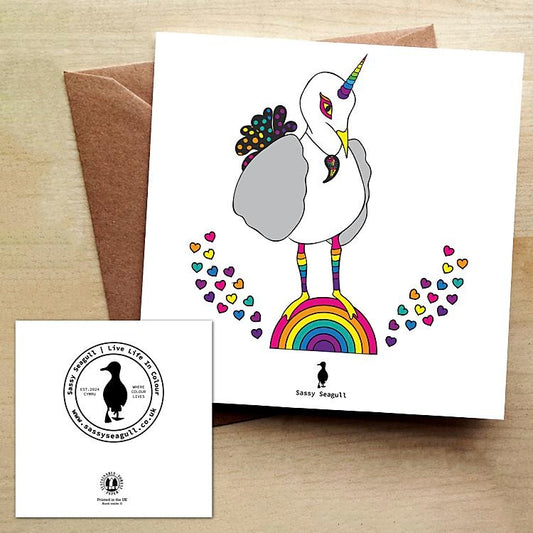 White greeting card with a rainbow unicorn seagull standing on a rainbow with rainbow coloured hearts. Card is on a brown envelope on a brown table.