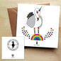 White greeting card with a rainbow unicorn seagull standing on a rainbow with rainbow coloured hearts. Card is on a brown envelope on a brown table.