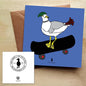 Blue greeting card featuring an illustrated seagull on a skateboard, with a brown envelope on a wooden table.